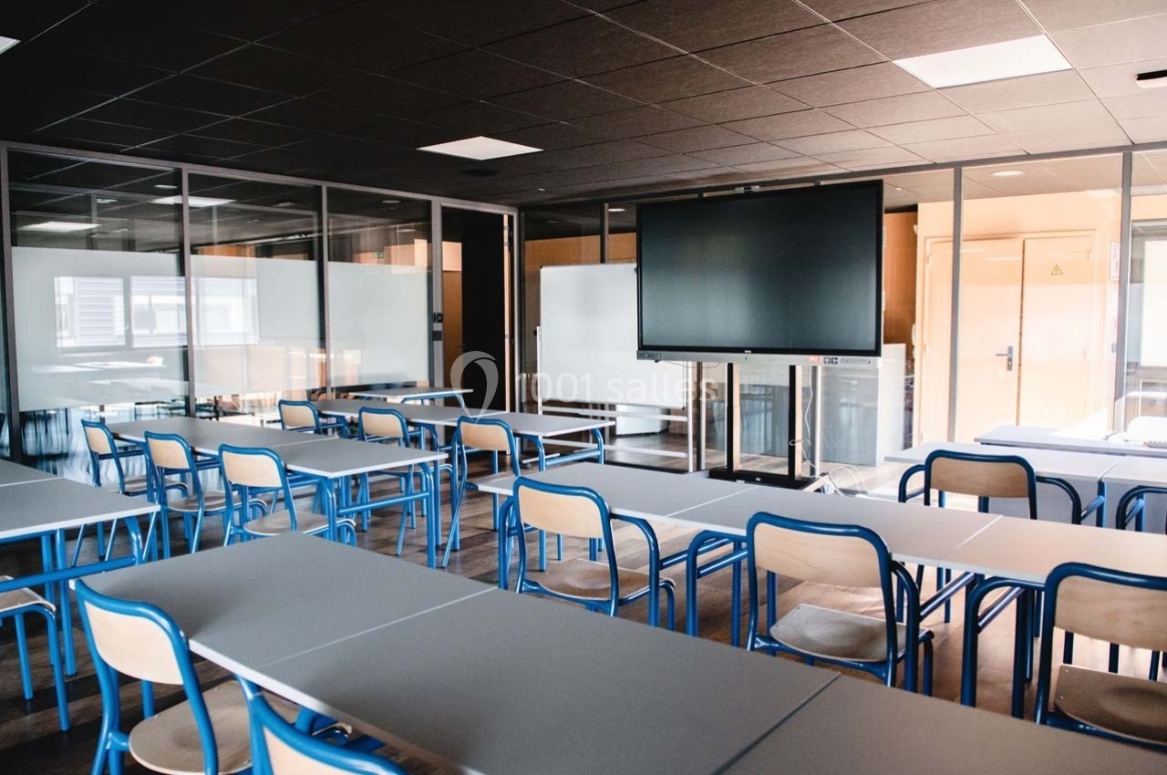Salle de classe moderne avec tables, chaises bleues, tableau blanc et écran numérique, éclairée par une lumière naturelle.