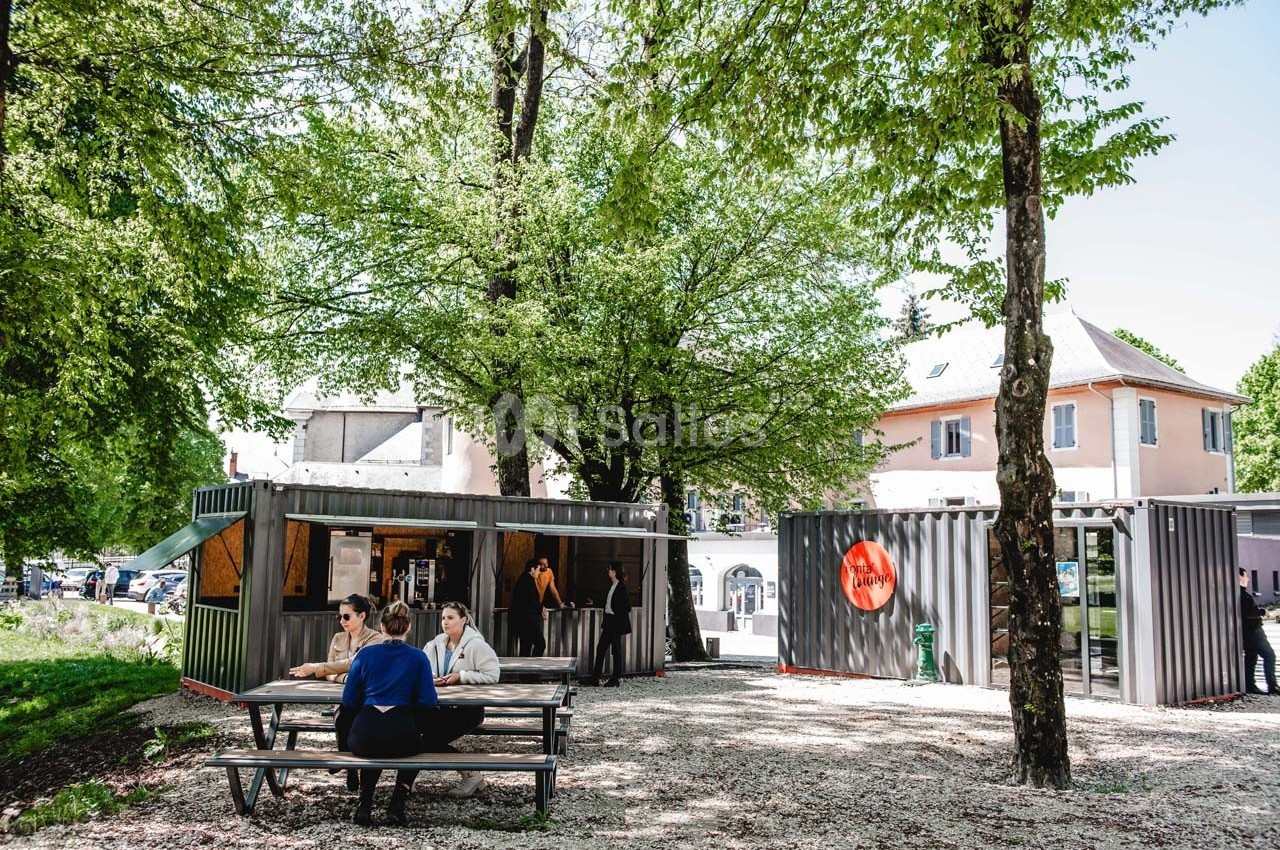 Des personnes discutent à une table de pique-nique près de conteneurs aménagés, entourés d'arbres et de bâtiments.