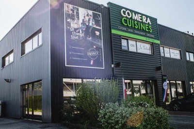 Façade d'un bâtiment commercial avec enseigne ’Comera Cuisines’ et affiches publicitaires, entouré de végétation.