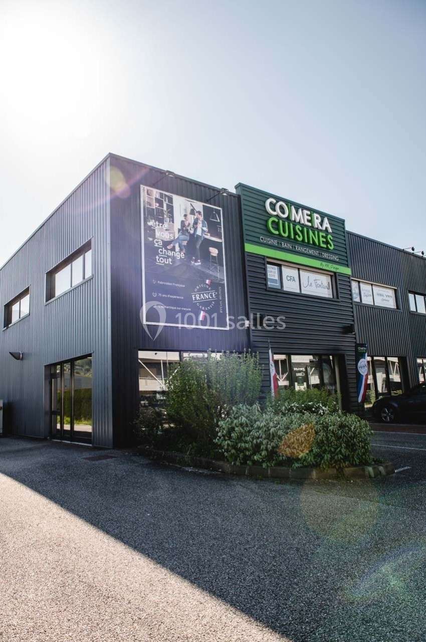 Façade d'un bâtiment commercial avec enseigne ’Comera Cuisines’ et affiches publicitaires, entouré de végétation.