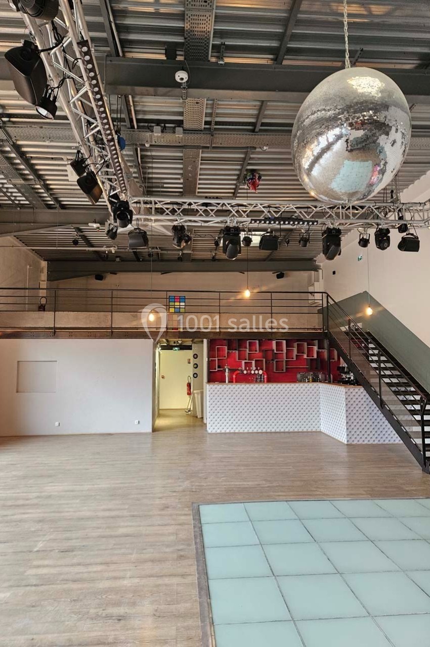 Salle spacieuse avec parquet, piste de danse vitrée, mezzanine, bar rouge et grande boule à facettes suspendue.