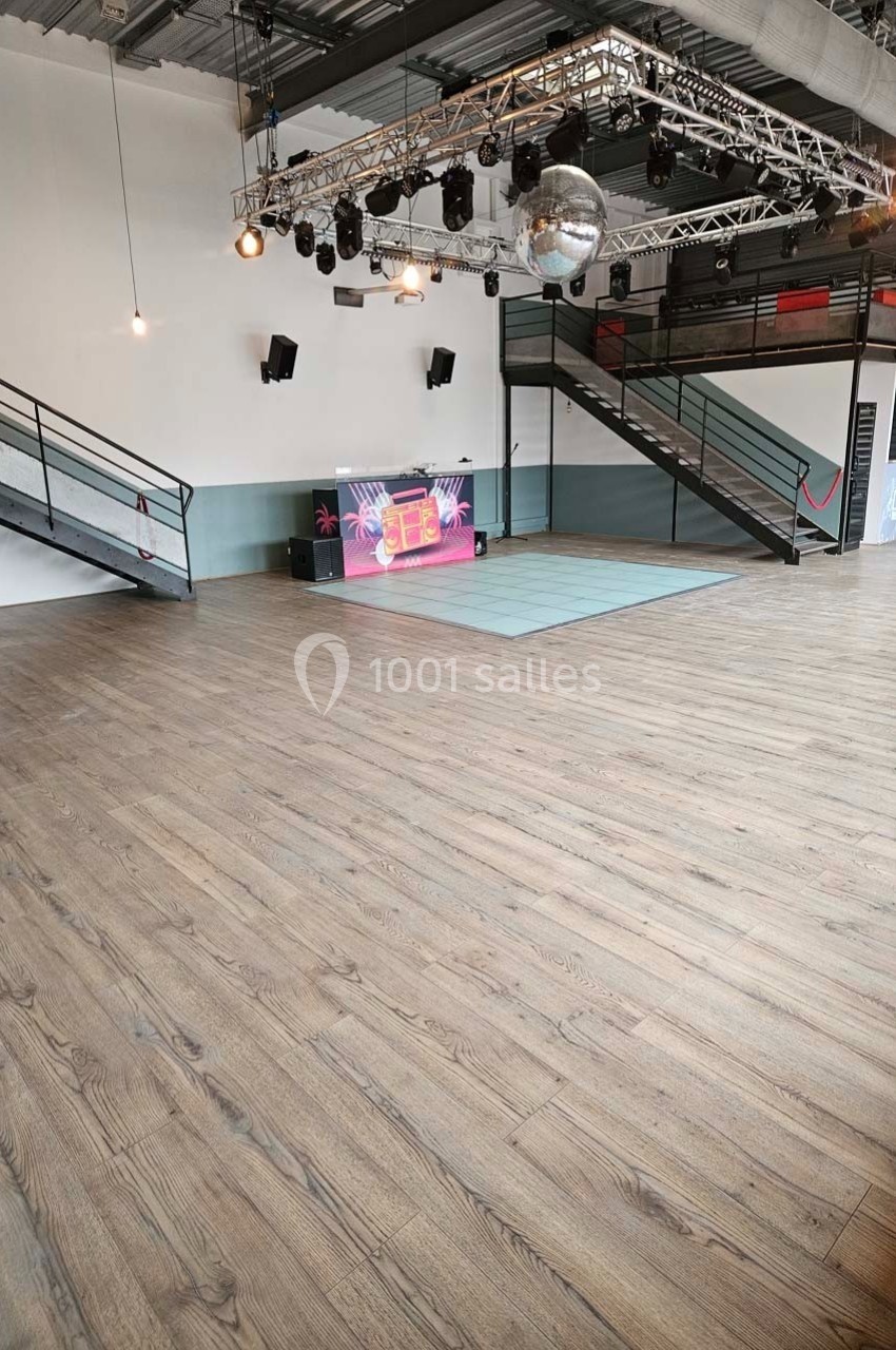 Salle spacieuse avec parquet, scène équipée d'un écran lumineux, éclairages suspendus et escaliers métalliques.