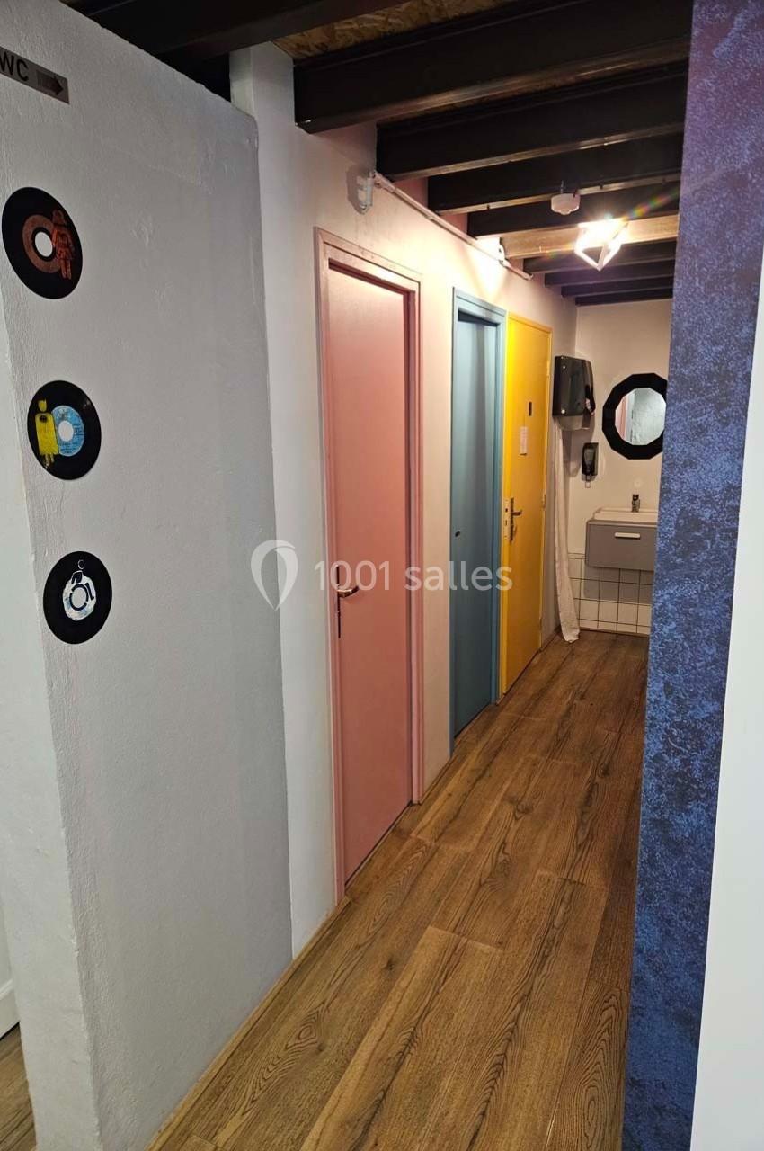 Couloir avec parquet, trois portes colorées (rose, bleu, jaune) et un lavabo avec miroir au fond.