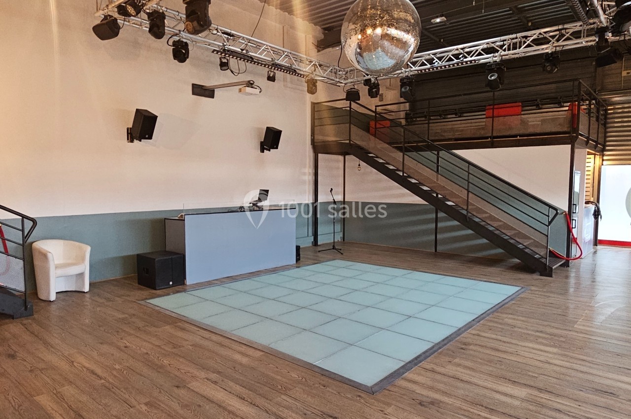 Salle vide avec piste de danse éclairée, cabine DJ, escalier métallique et boule à facettes suspendue.
