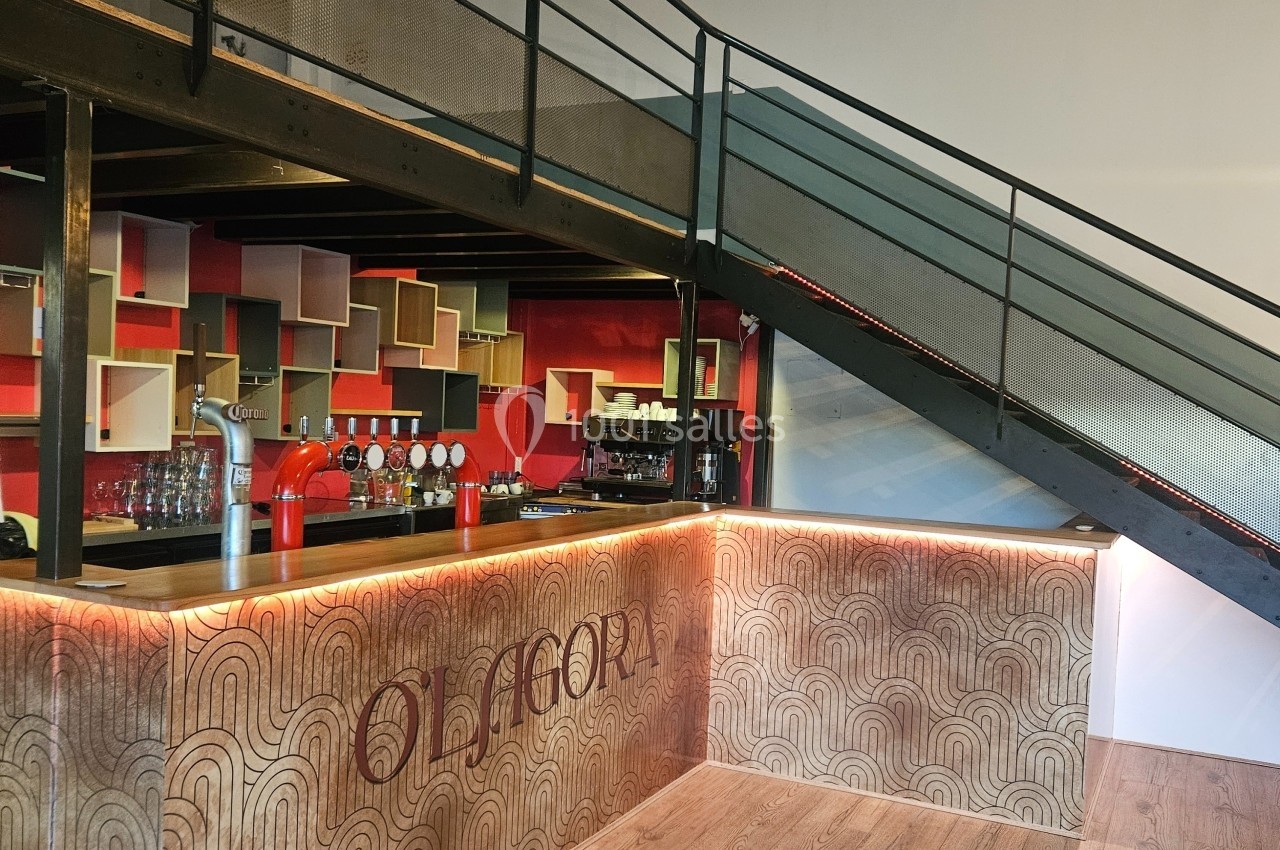 Bar moderne avec comptoir en bois gravé, étagères colorées en arrière-plan et escalier métallique sur le côté.