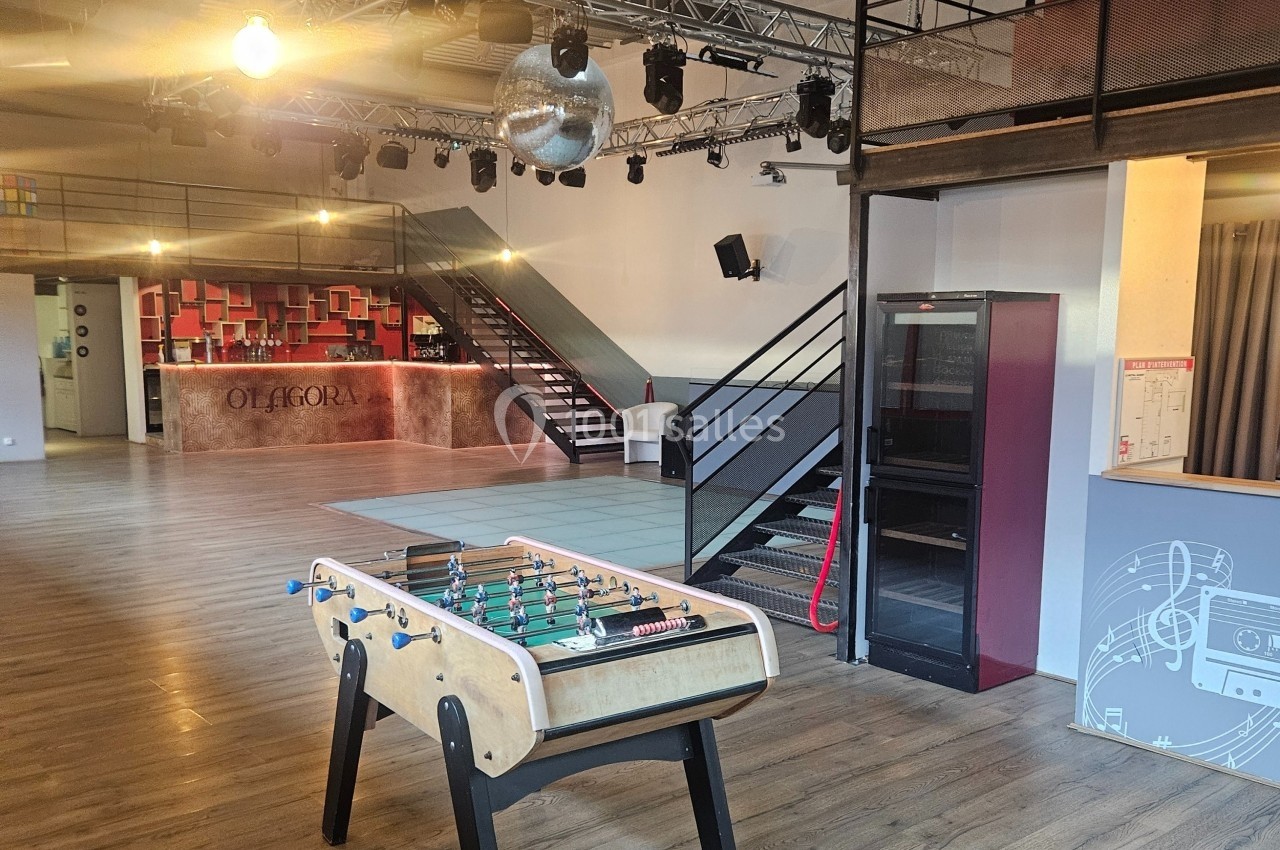 Salle lumineuse avec baby-foot au premier plan, escalier métallique, piste de danse et comptoir en arrière-plan.
