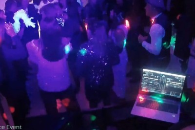 Un DJ en chemise rouge se tient derrière une table de mixage avec des lumières colorées en arrière-plan.