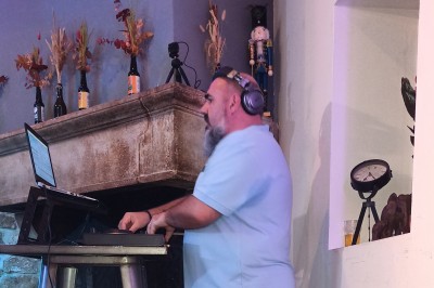 Un DJ en chemise rouge se tient derrière une table de mixage avec des lumières colorées en arrière-plan.