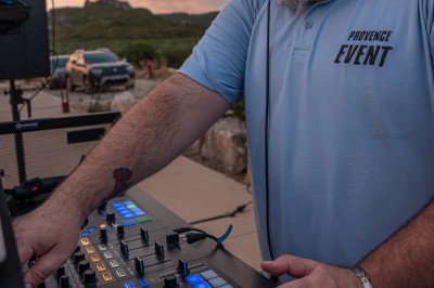 Un DJ en chemise rouge se tient derrière une table de mixage avec des lumières colorées en arrière-plan.