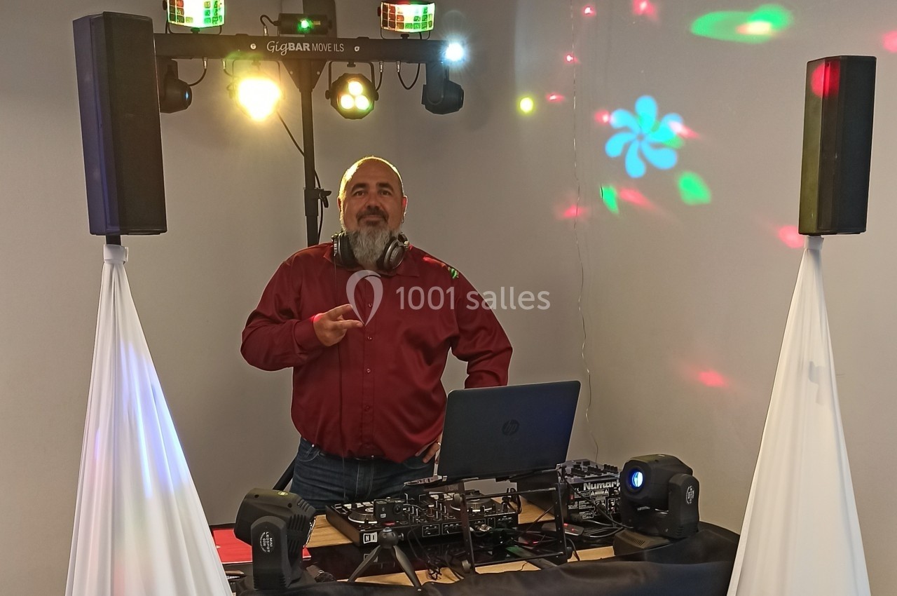 Un DJ en chemise rouge se tient derrière une table de mixage avec des lumières colorées en arrière-plan.