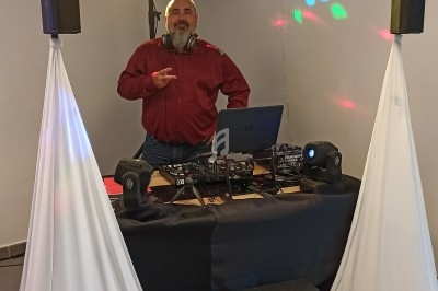 Un DJ en chemise rouge se tient derrière une table équipée de matériel audio et d'éclairage dans une salle.