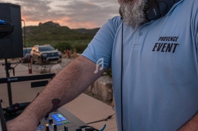 Un DJ mixant en plein air au coucher du soleil, avec une table de mixage et un casque autour du cou.
