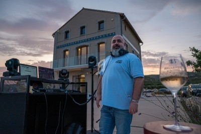 Un homme souriant devant un équipement de DJ, avec une maison et un ciel au coucher du soleil en arrière-plan.