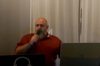 Homme en chemise rouge parlant dans un micro derrière une table avec des équipements audio et un ordinateur portable.