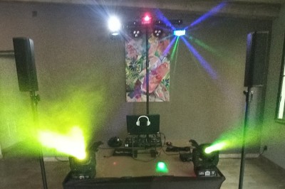 Un DJ en chemise rouge mixe de la musique sur une table équipée, avec des lumières et des haut-parleurs en arrière-plan.