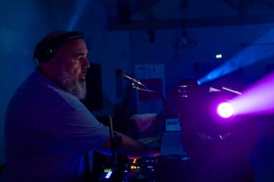 Un DJ en chemise rouge mixe de la musique sur une table équipée, avec des lumières et des haut-parleurs en arrière-plan.