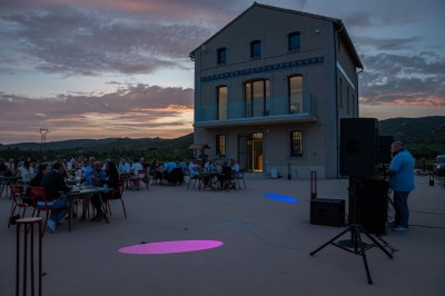 Un DJ en chemise rouge mixe de la musique sur une table équipée, avec des lumières et des haut-parleurs en arrière-plan.