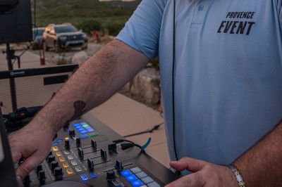 Un DJ en chemise rouge mixe de la musique sur une table équipée, avec des lumières et des haut-parleurs en arrière-plan.