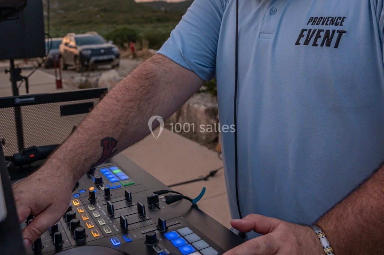 Un DJ en polo bleu manipule une table de mixage en extérieur, avec une voiture et un paysage en arrière-plan.