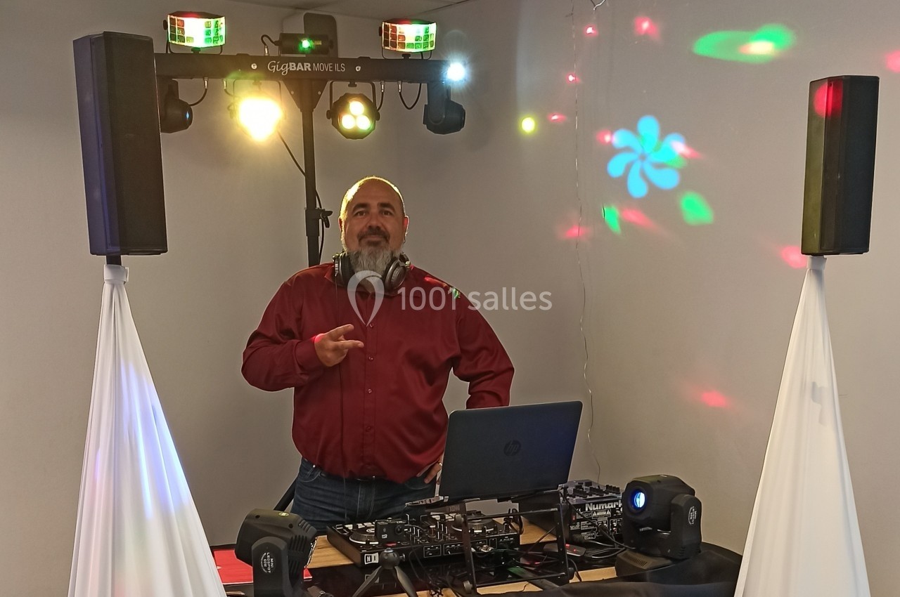 Un DJ en chemise rouge mixe de la musique sur une table équipée, avec des lumières et des haut-parleurs en arrière-plan.