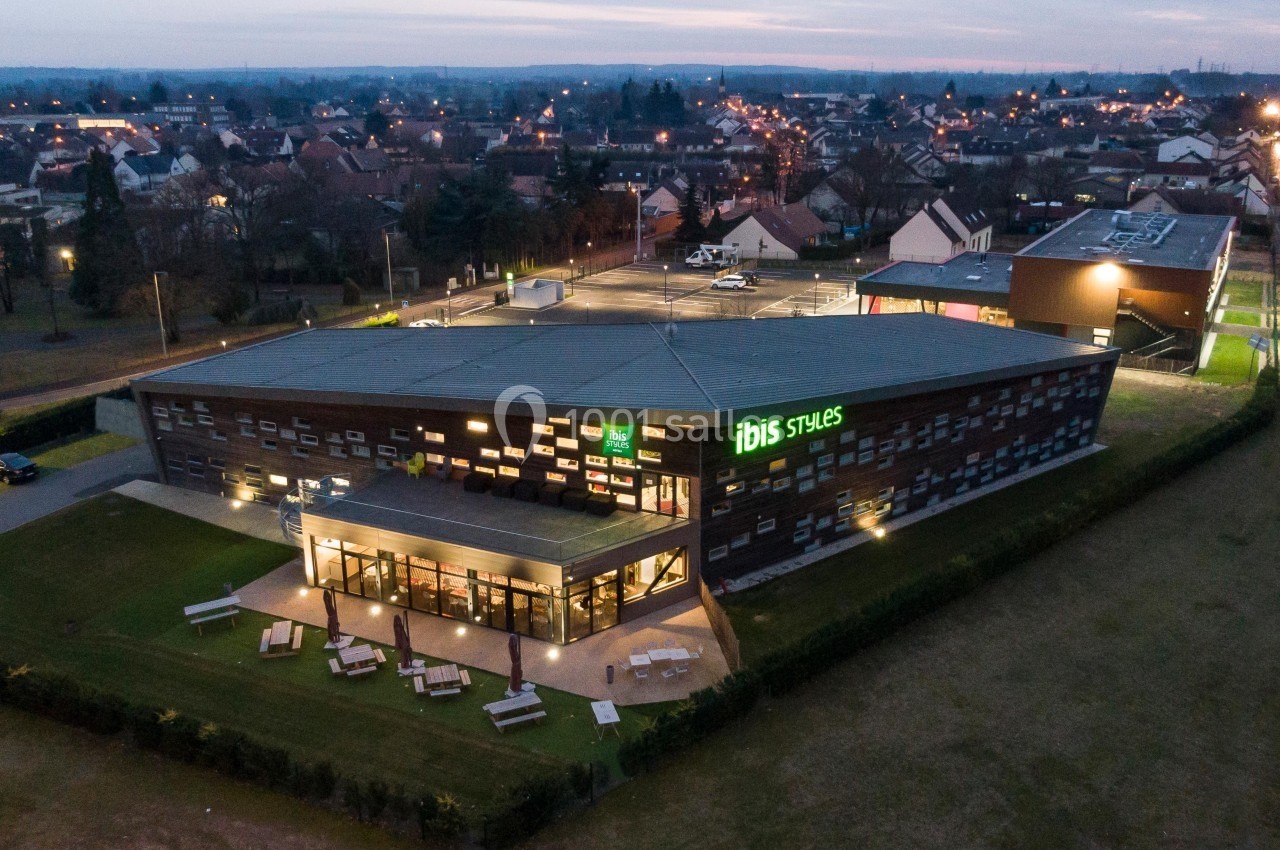 Vue aérienne d'un hôtel Ibis Styles moderne entouré de verdure, avec un parking et des habitations en arrière-plan.