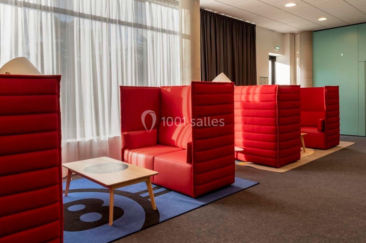 Fauteuils rouges avec cloisons hautes disposés dans un espace lumineux, près de tables basses et de grandes fenêtres.