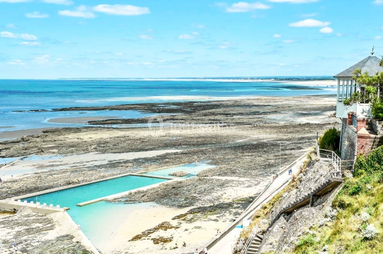 Vue d'une piscine d'eau de mer surplombant une plage rocheuse et l'océan sous un ciel bleu dégagé.