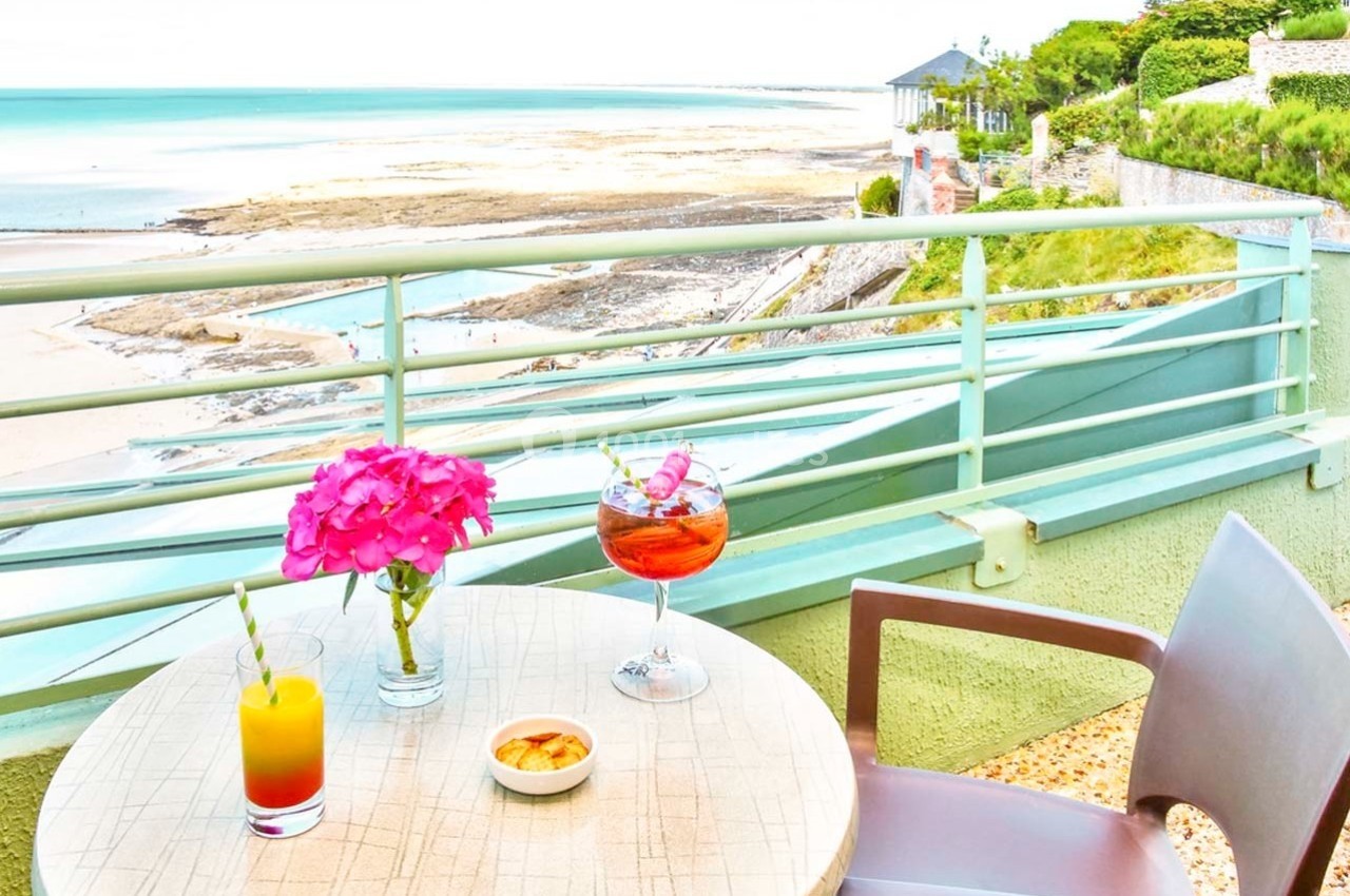 Vue sur une plage depuis une terrasse avec table, boissons, fleurs roses et fauteuils, entourée de verdure.