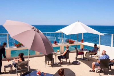 Façade d'un bâtiment clair avec terrasse, escalier en pierre et mobilier extérieur sous un ciel dégagé.