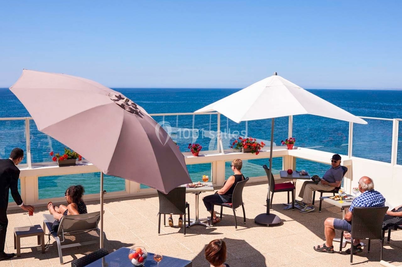 Terrasse ensoleillée avec vue sur la mer, des personnes assises sous des parasols autour de tables.