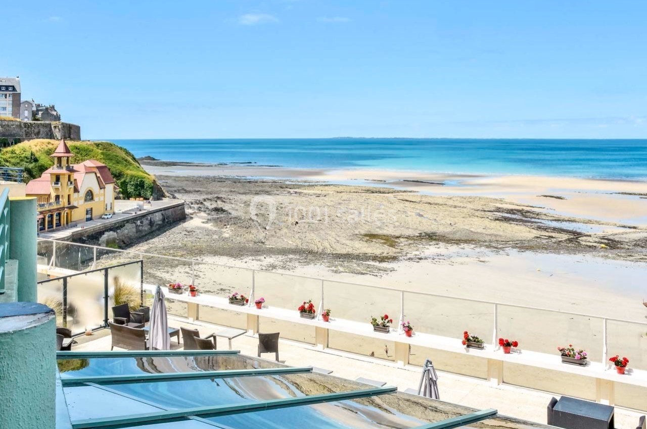 Vue sur une plage de sable à marée basse avec des bâtiments en bord de mer et une terrasse ornée de fleurs.