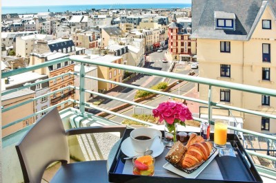 Façade d'un bâtiment clair avec terrasse, escalier en pierre et mobilier extérieur sous un ciel dégagé.