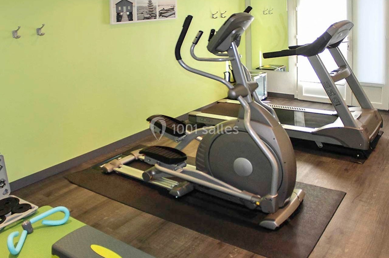 Salle de sport équipée d'un vélo elliptique, d'un tapis de course et d'accessoires sur un sol en bois.