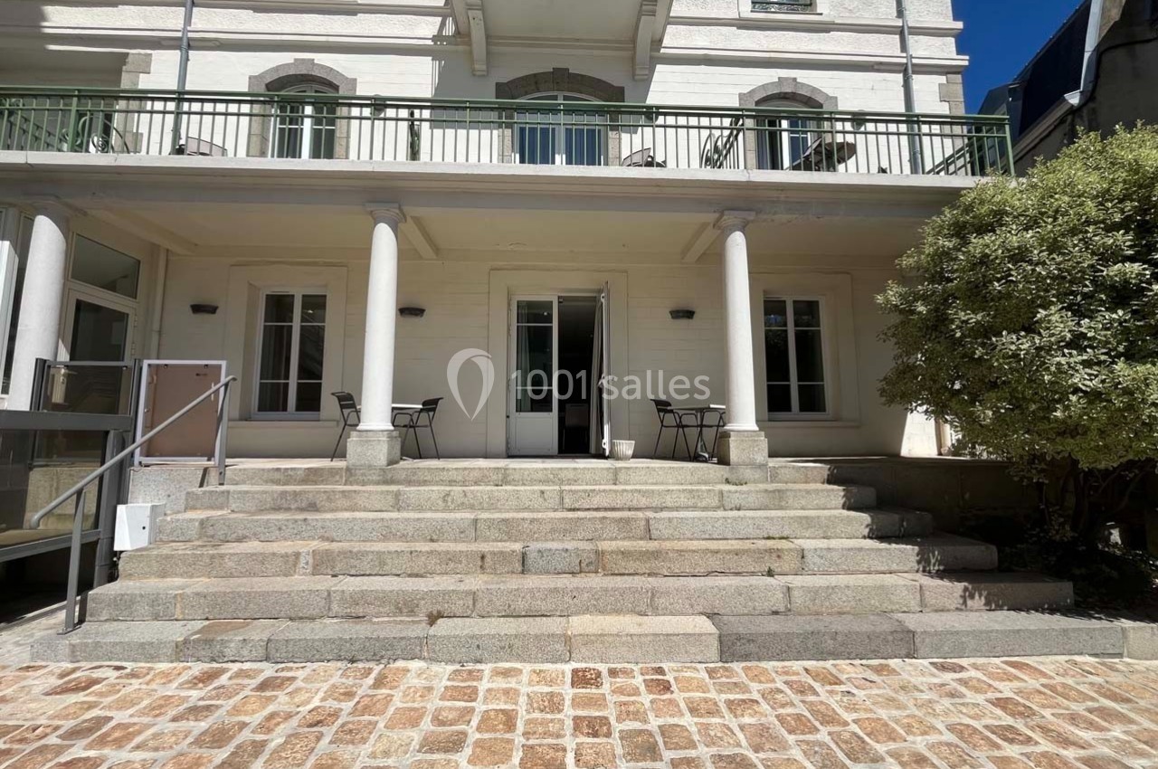 Façade d'un bâtiment clair avec terrasse, escalier en pierre et mobilier extérieur sous un ciel dégagé.