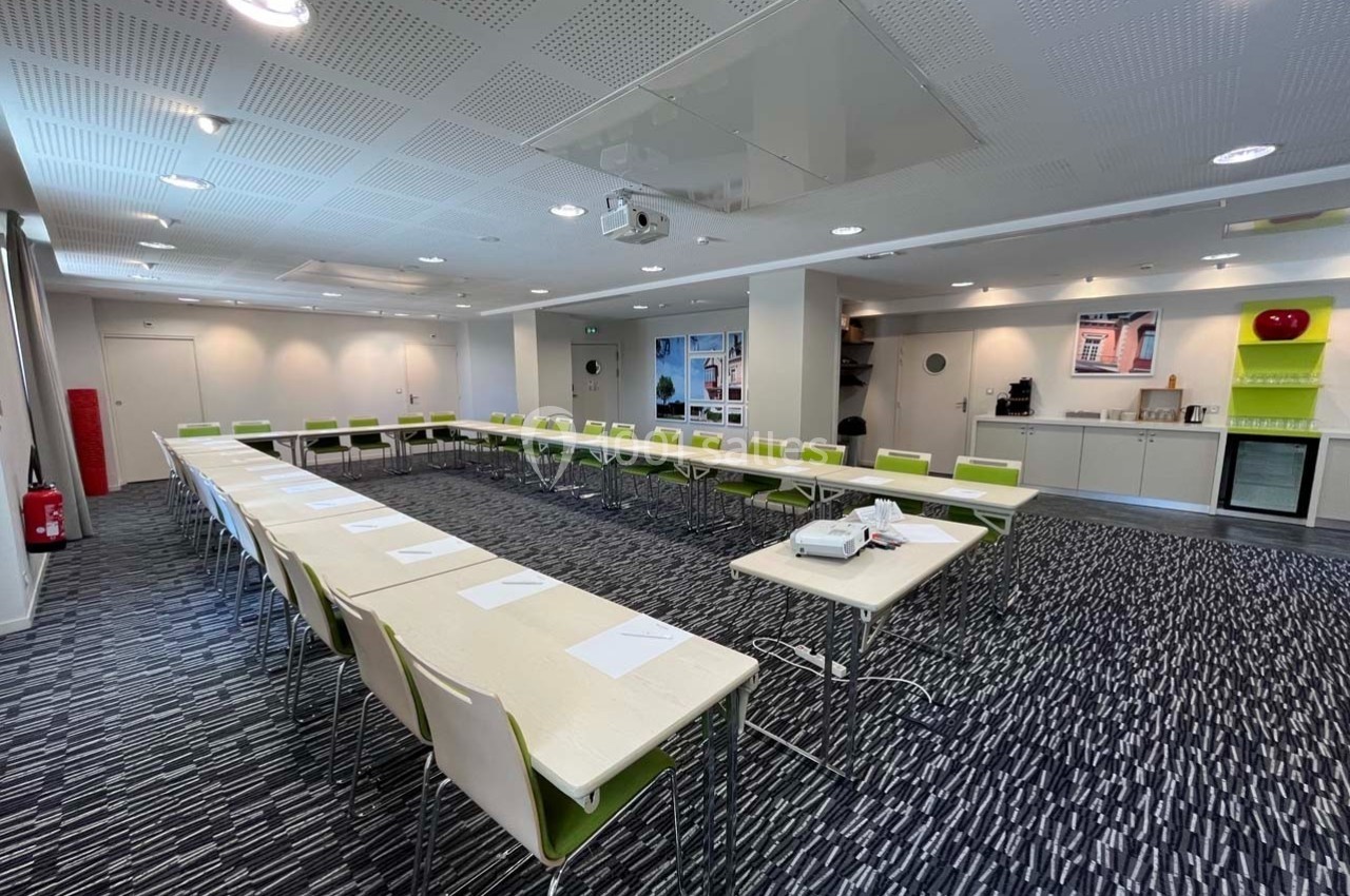 Salle de réunion lumineuse avec tables disposées en U, chaises blanches et vertes, et équipement audiovisuel.