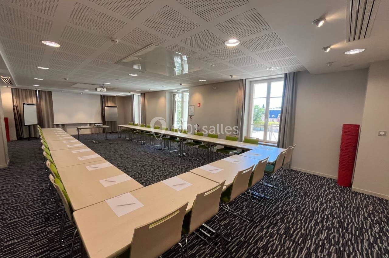 Salle de réunion lumineuse avec tables disposées en U, chaises vertes, écran de projection et fenêtres donnant sur l…