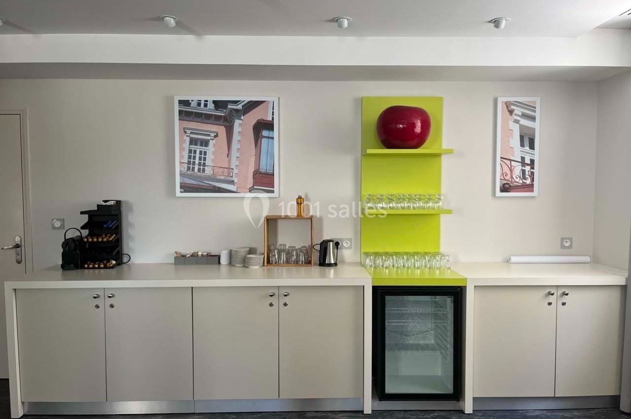 Coin café avec machine à café, tasses, verres, mini-réfrigérateur et décoration murale dans un espace lumineux.