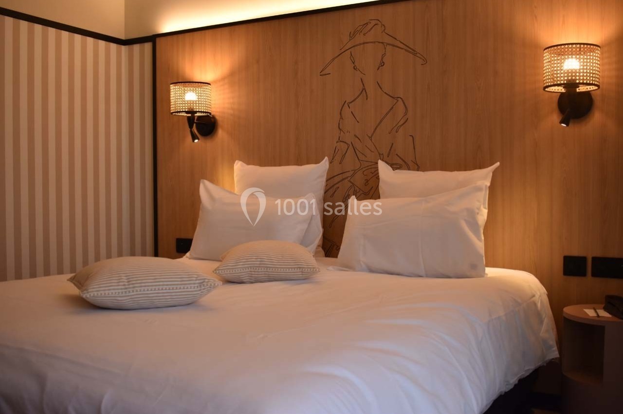 Lit double avec draps blancs, deux coussins décoratifs et éclairage mural dans une chambre à la décoration sobre.