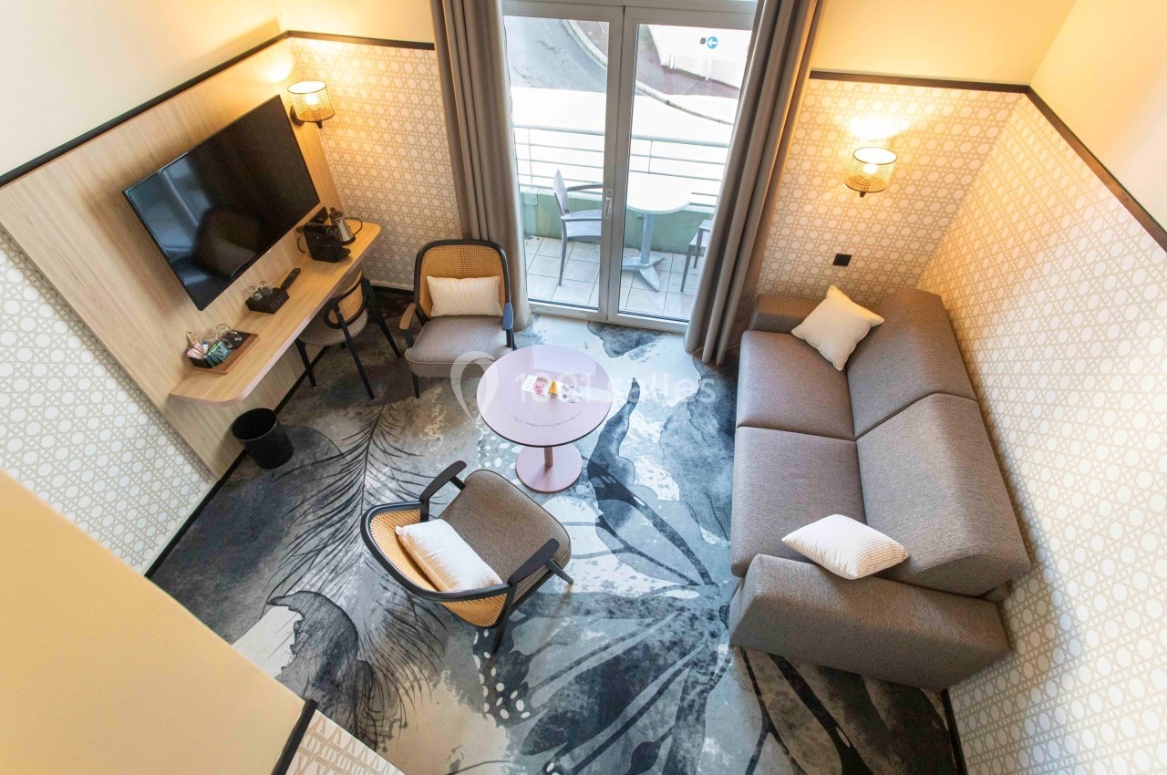 Salon moderne avec canapé, fauteuils, table basse, télévision murale et accès à un balcon avec mobilier extérieur.