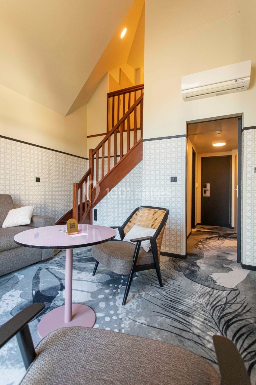 Salon d'un intérieur moderne avec fauteuils, table basse rose, escalier en bois et éclairage chaleureux.