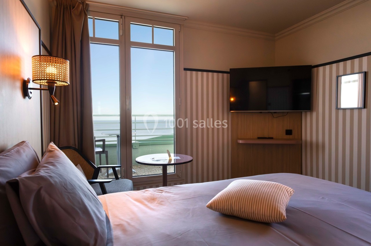 Chambre d'hôtel lumineuse avec lit, télévision, table et vue sur la mer à travers une grande baie vitrée.