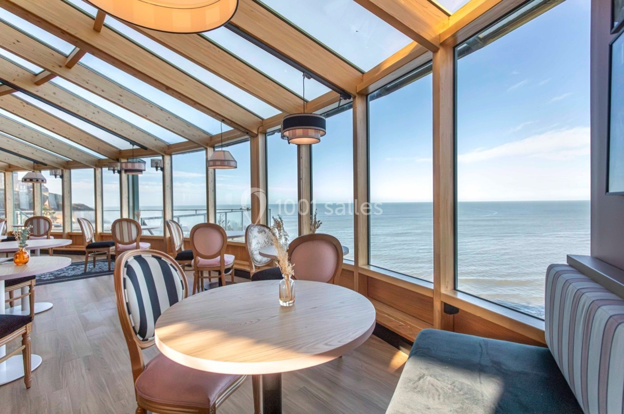 Salle lumineuse avec grandes baies vitrées offrant une vue panoramique sur la mer, tables et chaises en bois clair.