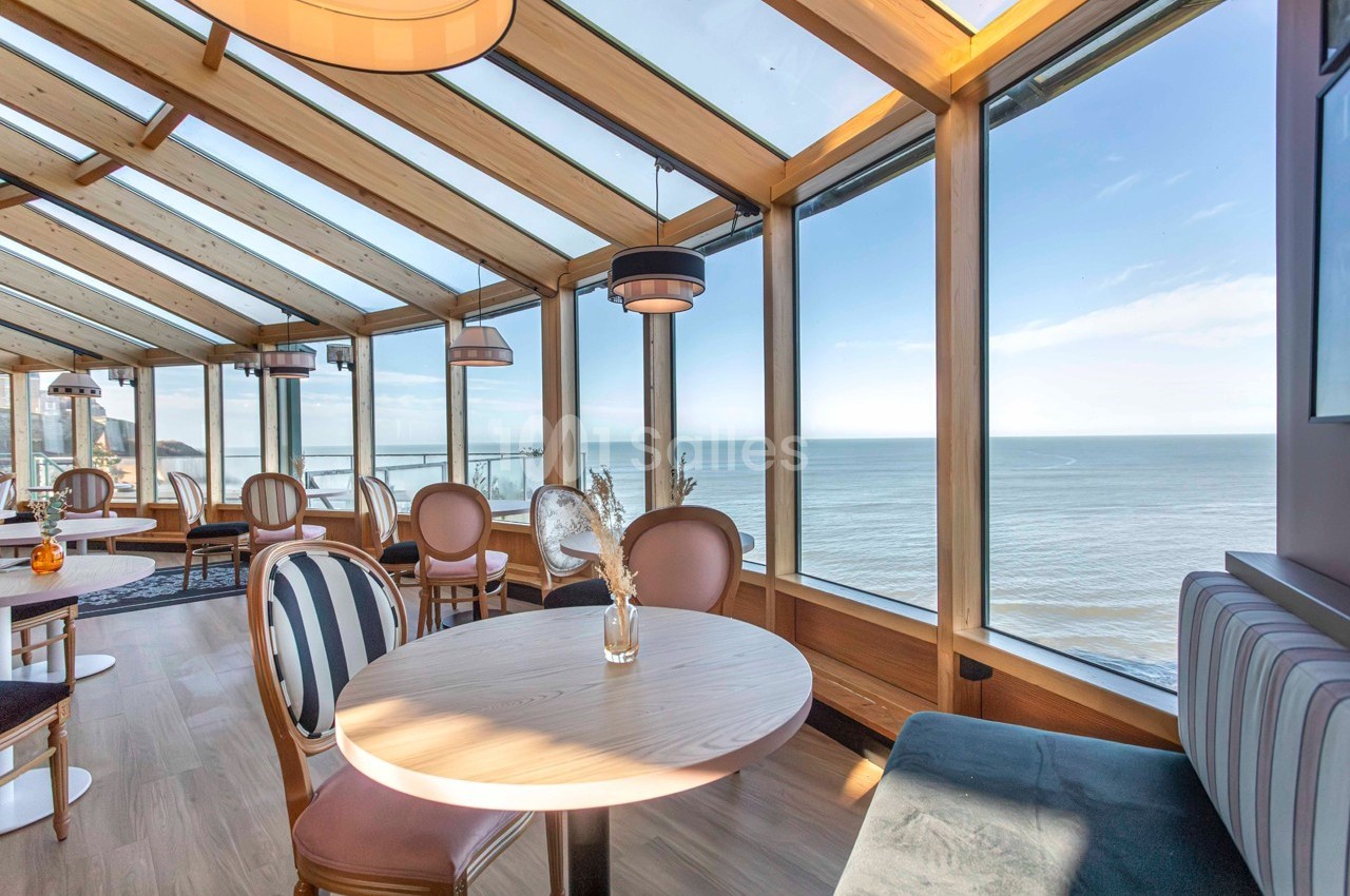 Salle lumineuse avec grandes baies vitrées offrant une vue panoramique sur la mer, tables et chaises en bois clair.