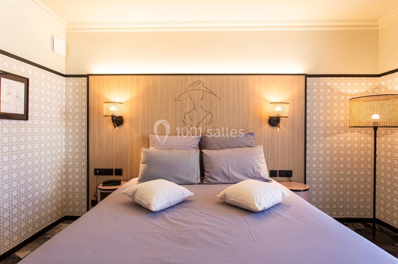 Lit double avec draps violets, deux lampes murales et une décoration murale en bois dans une chambre lumineuse.