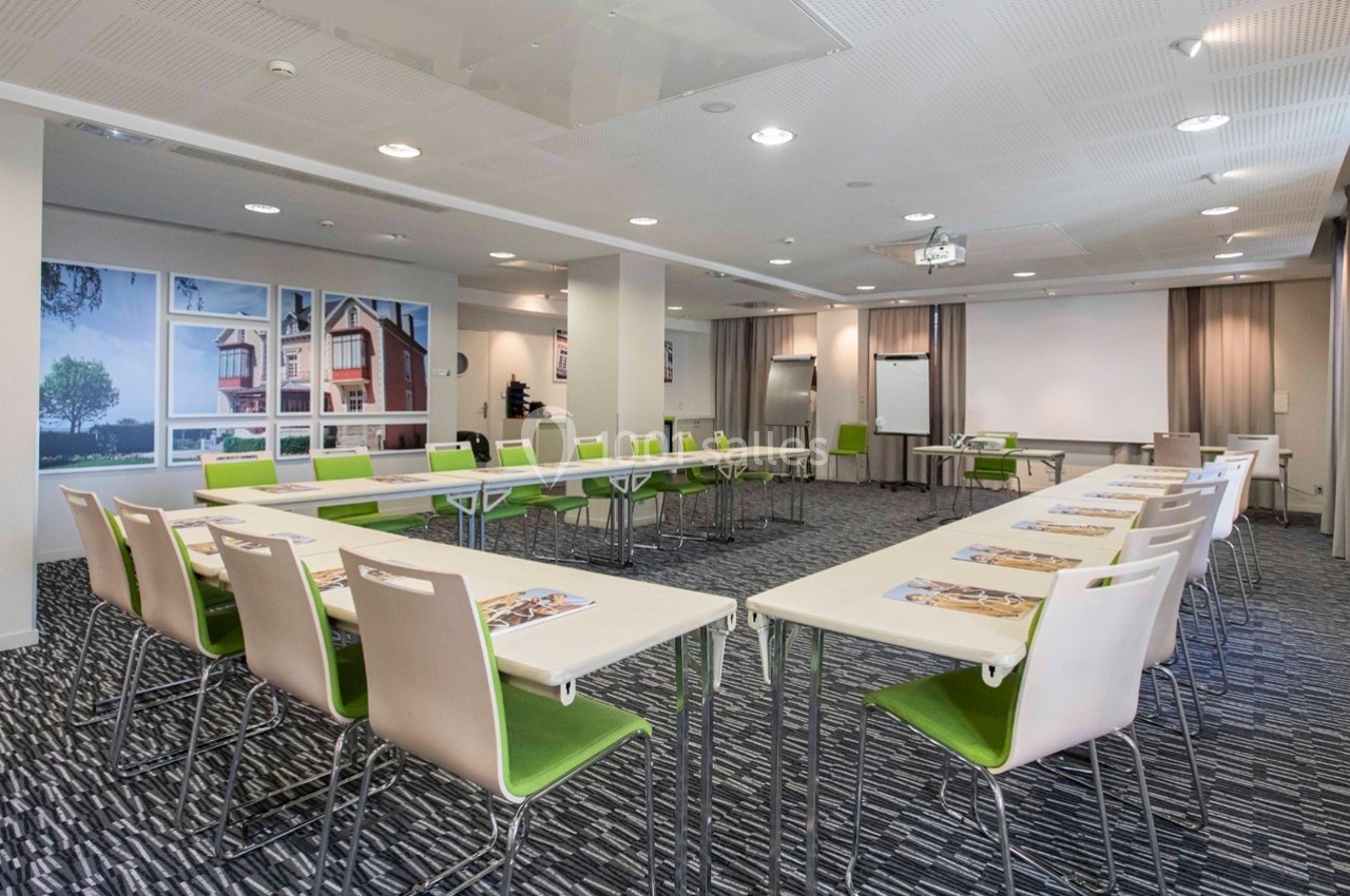 Salle de réunion lumineuse avec tables disposées en U, chaises vertes et écran de projection en fond.