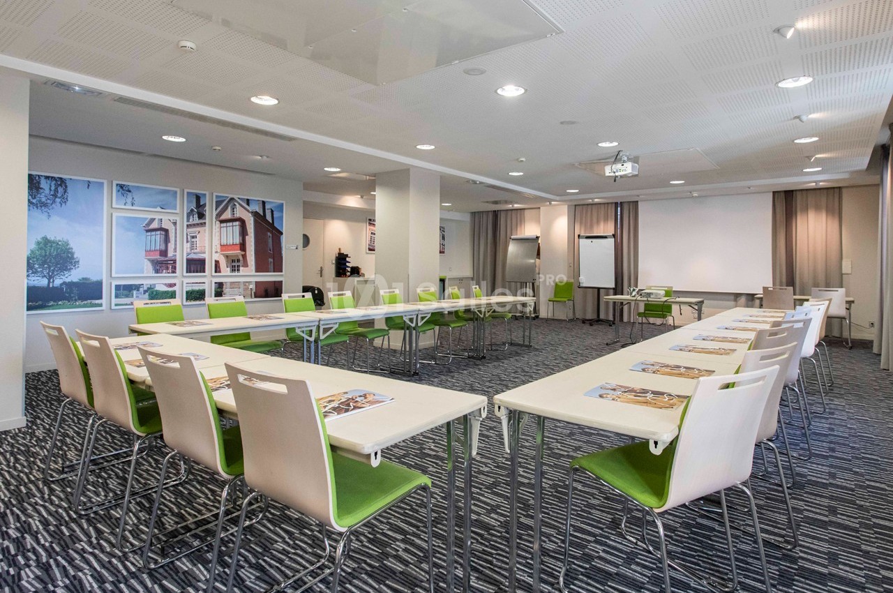 Salle de réunion lumineuse avec tables disposées en U, chaises vertes et écran de projection en fond.