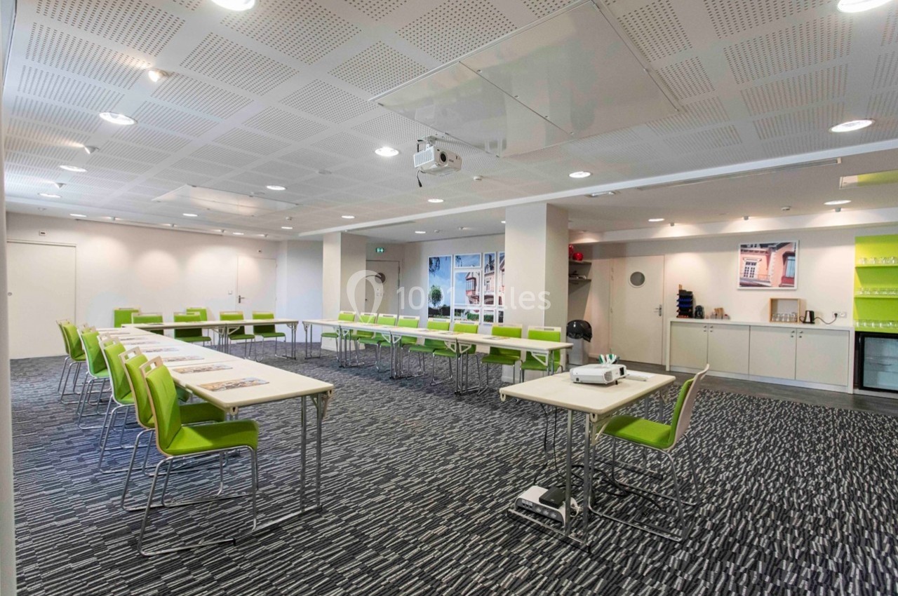 Salle de réunion lumineuse avec tables en U, chaises vertes, vidéoprojecteur et étagères en arrière-plan.