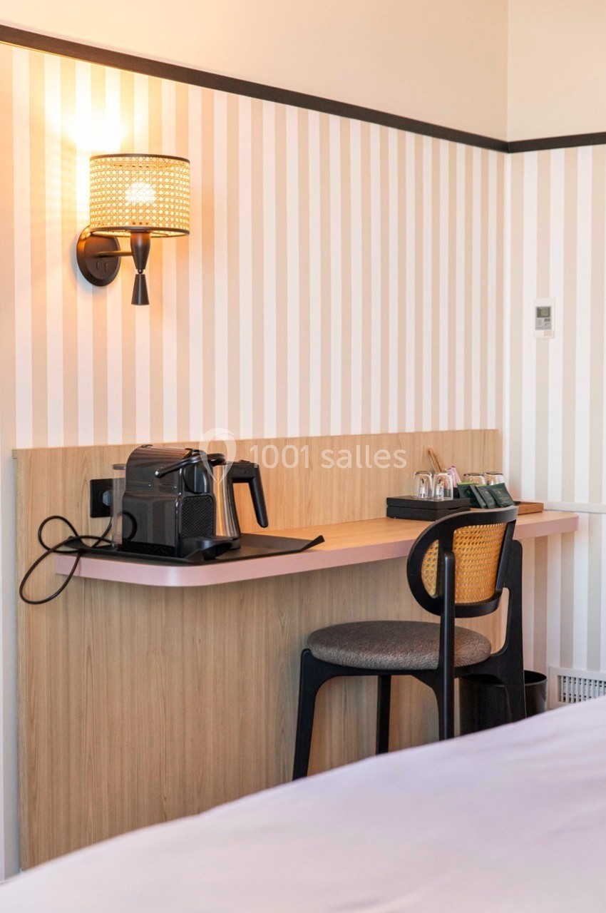 Bureau en bois clair avec une chaise, une machine à café, des tasses et une lampe murale allumée dans une chambre.