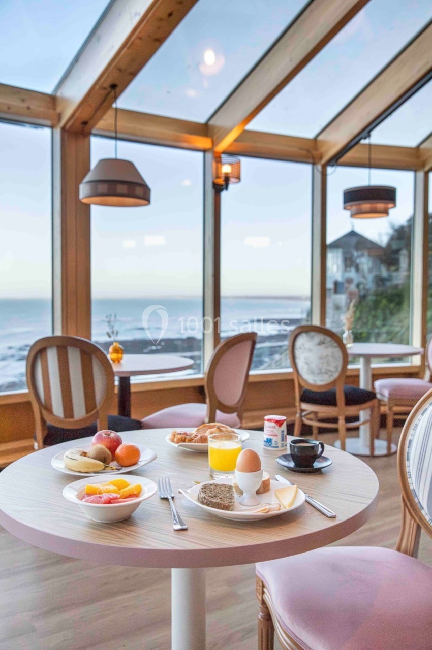 Petit-déjeuner servi sur une table ronde dans une salle lumineuse avec vue sur la mer à travers de grandes baies vitrées.
