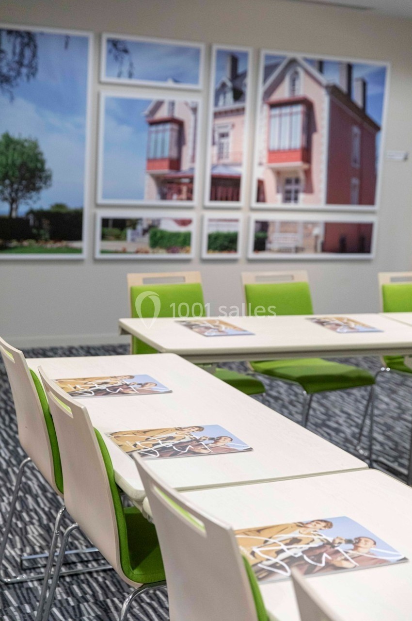 Salle de réunion avec des chaises vertes, des tables blanches et des brochures posées, décorée de photos de maisons.